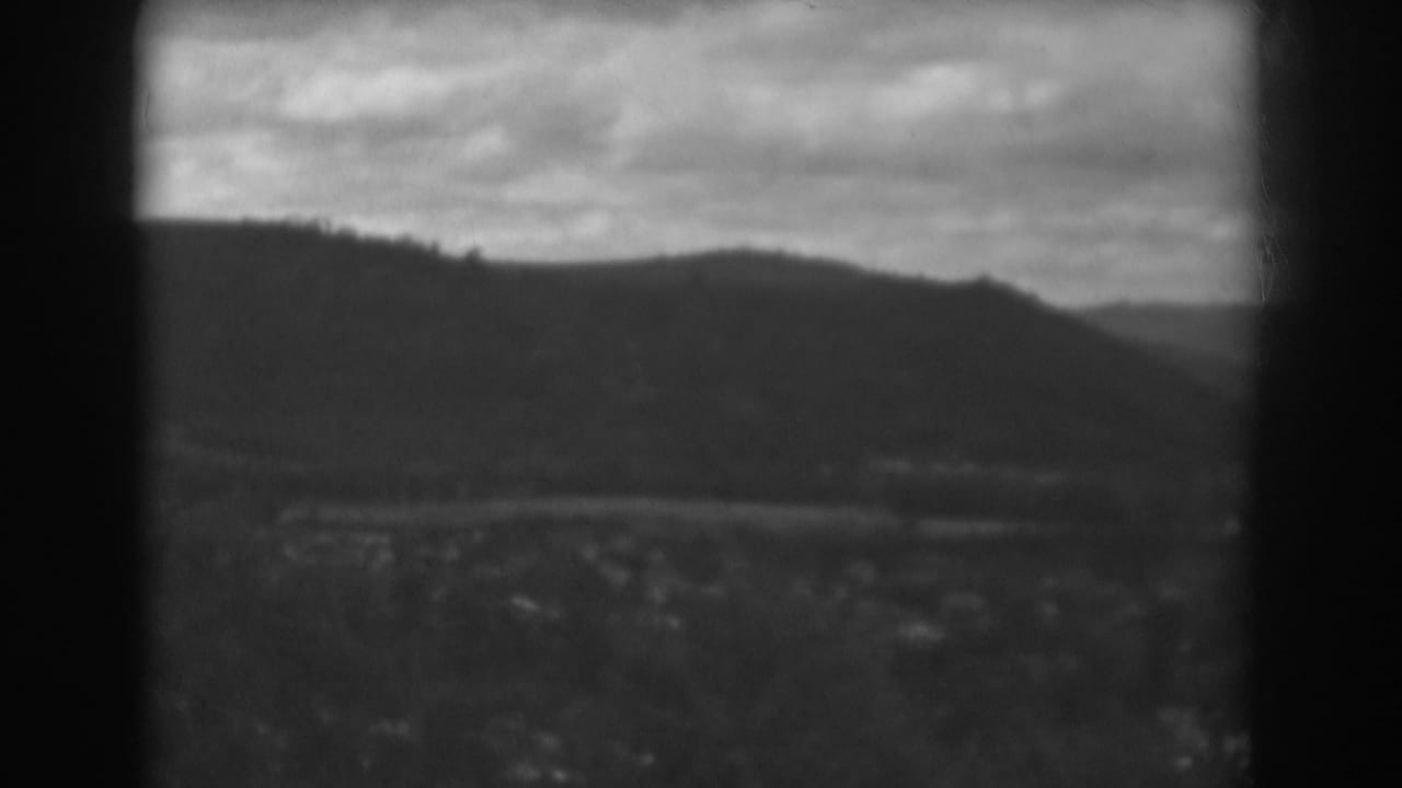 Dark window trim frames a distant valley, road, mountain ridge, cloudy sky, trees, and scattered buildings.