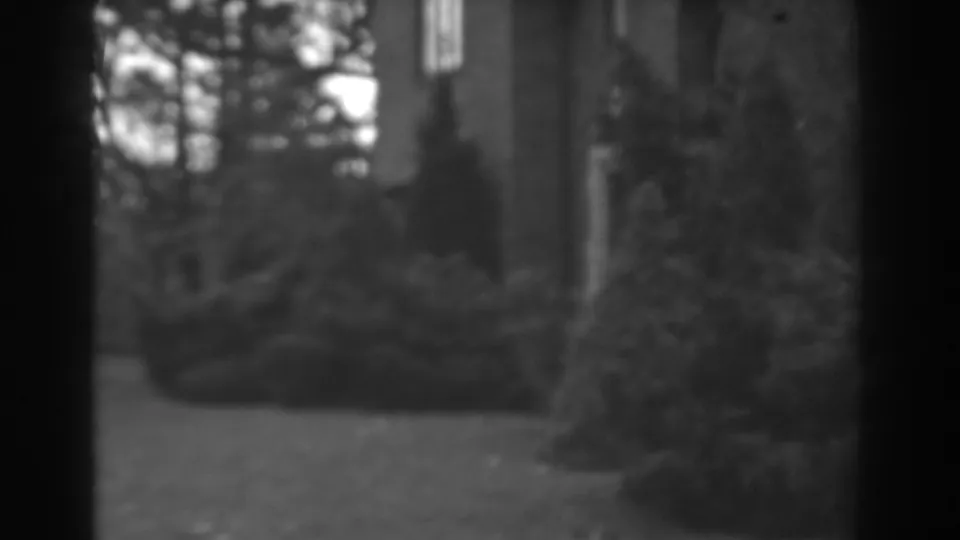 1938 Winchester, Maryland home-movie frame showing a large house facade behind evergreen shrubs and mature trees.