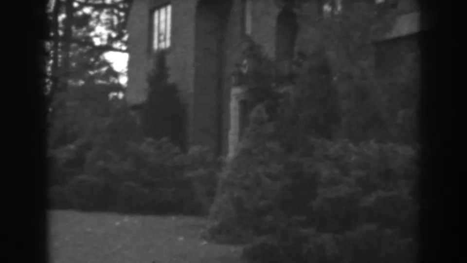 Black-and-white 8mm frame from Winchester, Maryland showing a house wall and window behind thick evergreen landscaping.