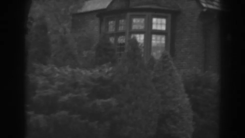 Static 8mm view of a Winchester brick house facade with bay windows and heavy shrubbery in the foreground.
