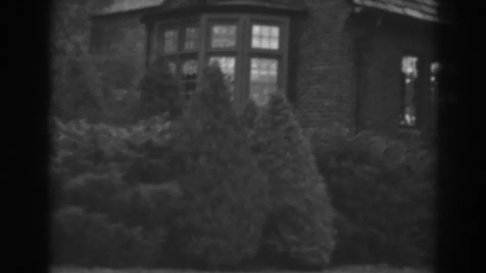 Home-movie frame of a brick residence in Winchester, with shrubs covering much of the lower facade and a darker doorway area at right.