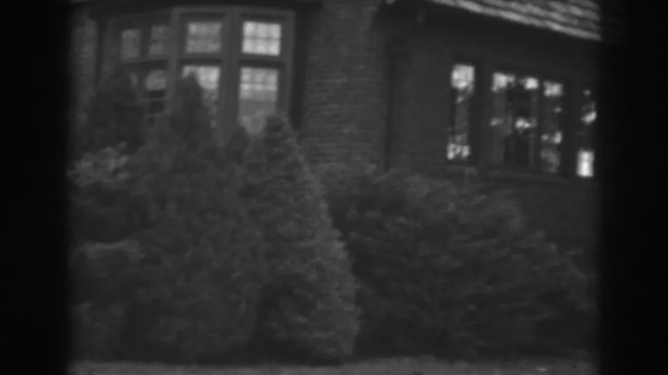 1938 8mm frame showing the Winchester brick house exterior, garden shrubs, bay window, and a shadowed entry or porch area.