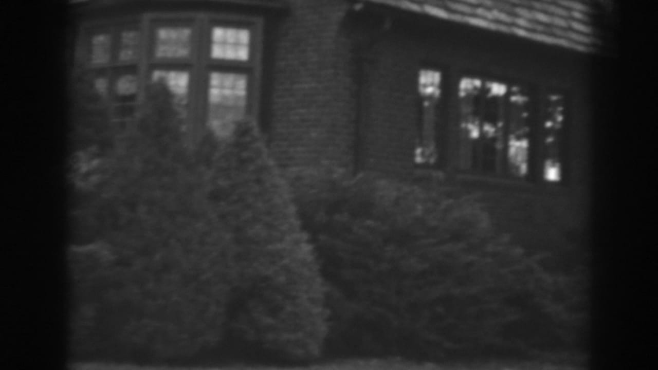 1938 Winchester, Maryland home-movie frame showing a brick house side, multi-pane windows, roofline, and trimmed shrubs.