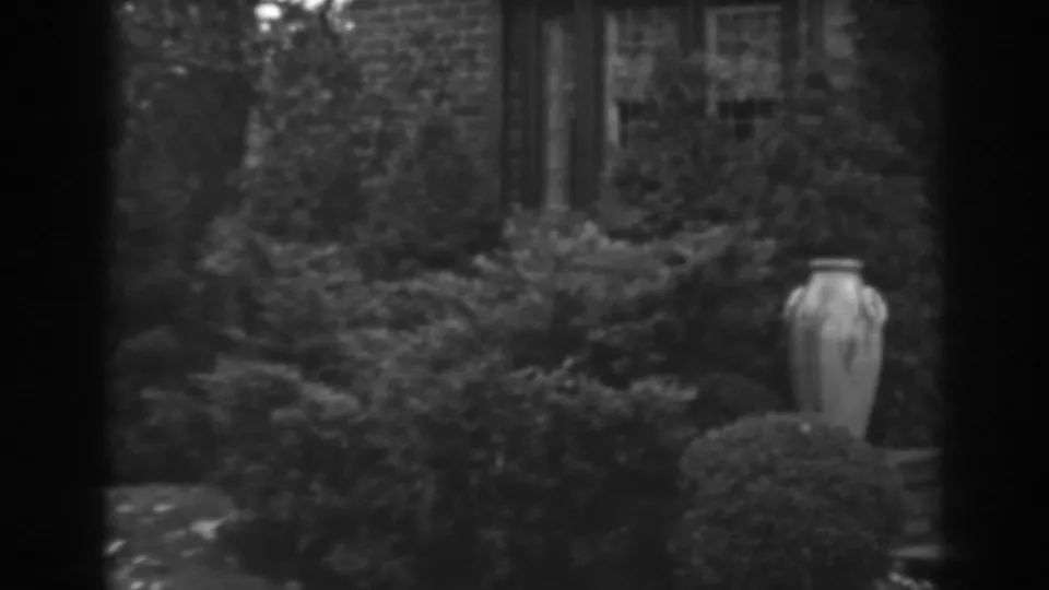 1938 Winchester, Maryland home-movie frame showing a stone garden urn, shrubs, and part of a brick house exterior.