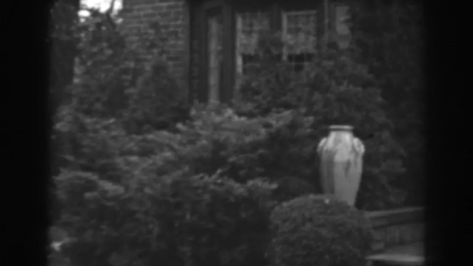 Black-and-white 8mm frame of a residential garden with a stone urn, shrubs, window, and brick exterior in 1938 Winchester, Maryland.