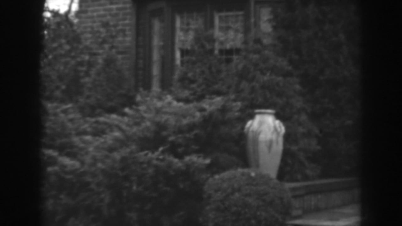 1938 Winchester, Maryland home-movie frame showing a pale garden urn, dense shrubs, brick wall, and multi-pane residential window.