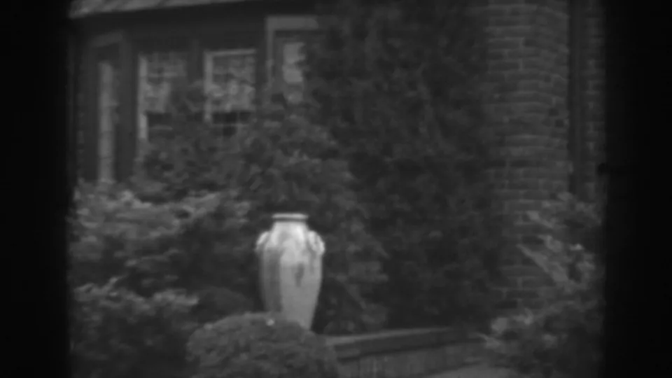 1938 Winchester, Maryland frame showing the urn lower in view, surrounded by plantings and backed by brickwork and residential windows.