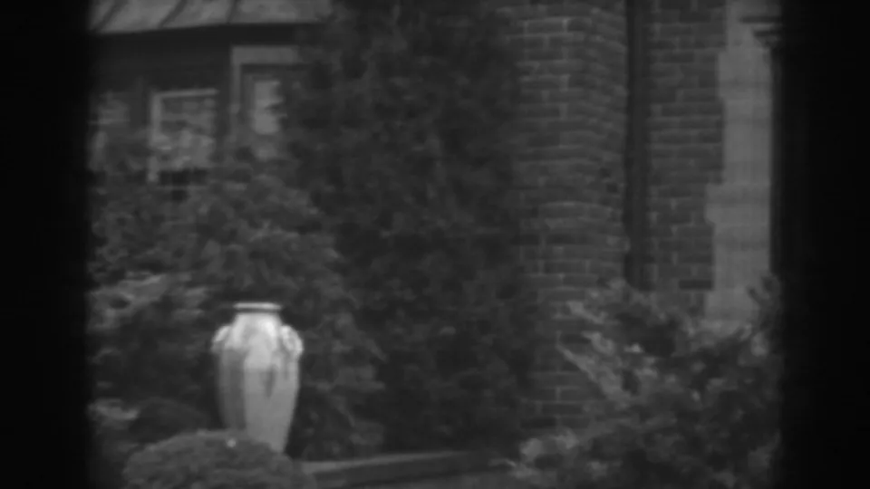 1938 Winchester, Maryland frame showing a pale urn below dense shrubs, brick wall, and a partial doorway or window edge at the right.