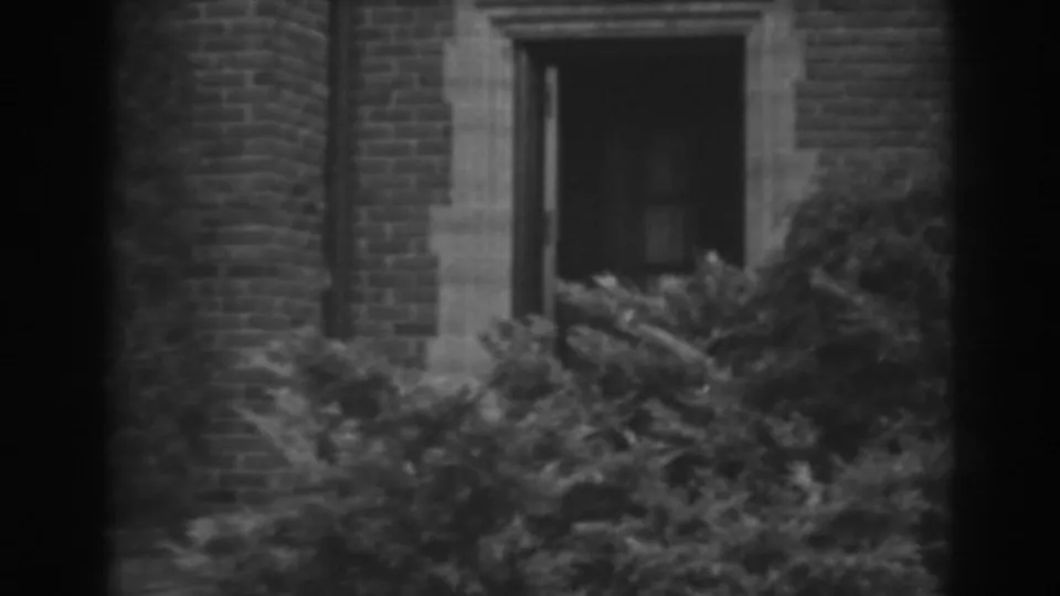 Soft black-and-white 8mm frame of a Winchester, Maryland brick facade with stone trim and heavy shrubbery in the foreground.