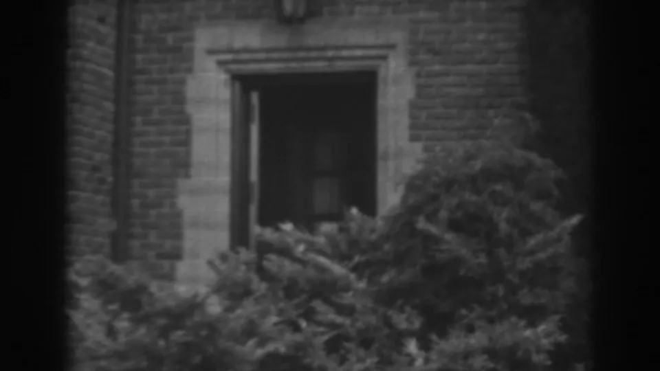 1938 8mm exterior in Winchester, Maryland, showing a brick building opening framed by stone and surrounded by dense bushes.