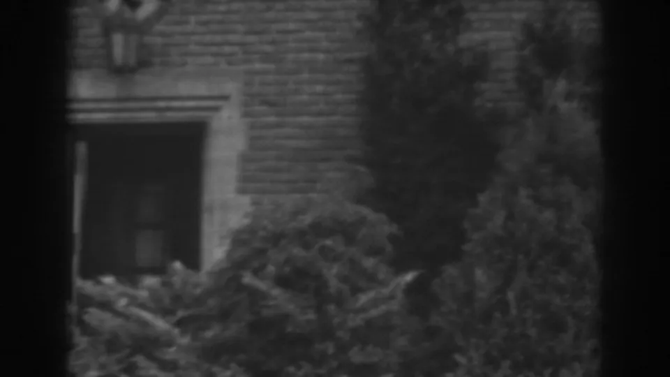 The camera holds on the brick house exterior, with a shadowed doorway, stone trim, shrubs, and dark vertical edges from the 8mm frame.