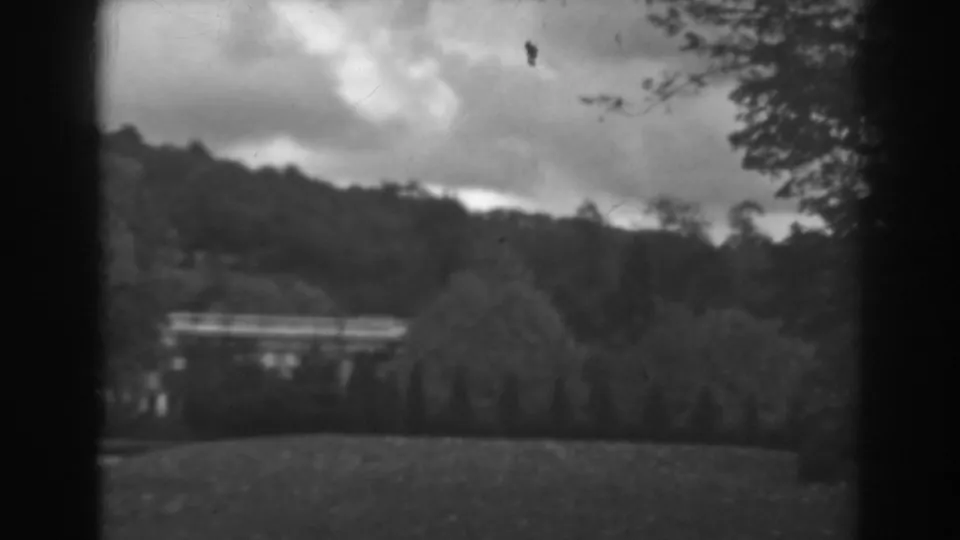 Black-and-white 8mm frame from the 1938 Winchester home movie, looking across grass and trees toward a distant hillside structure.