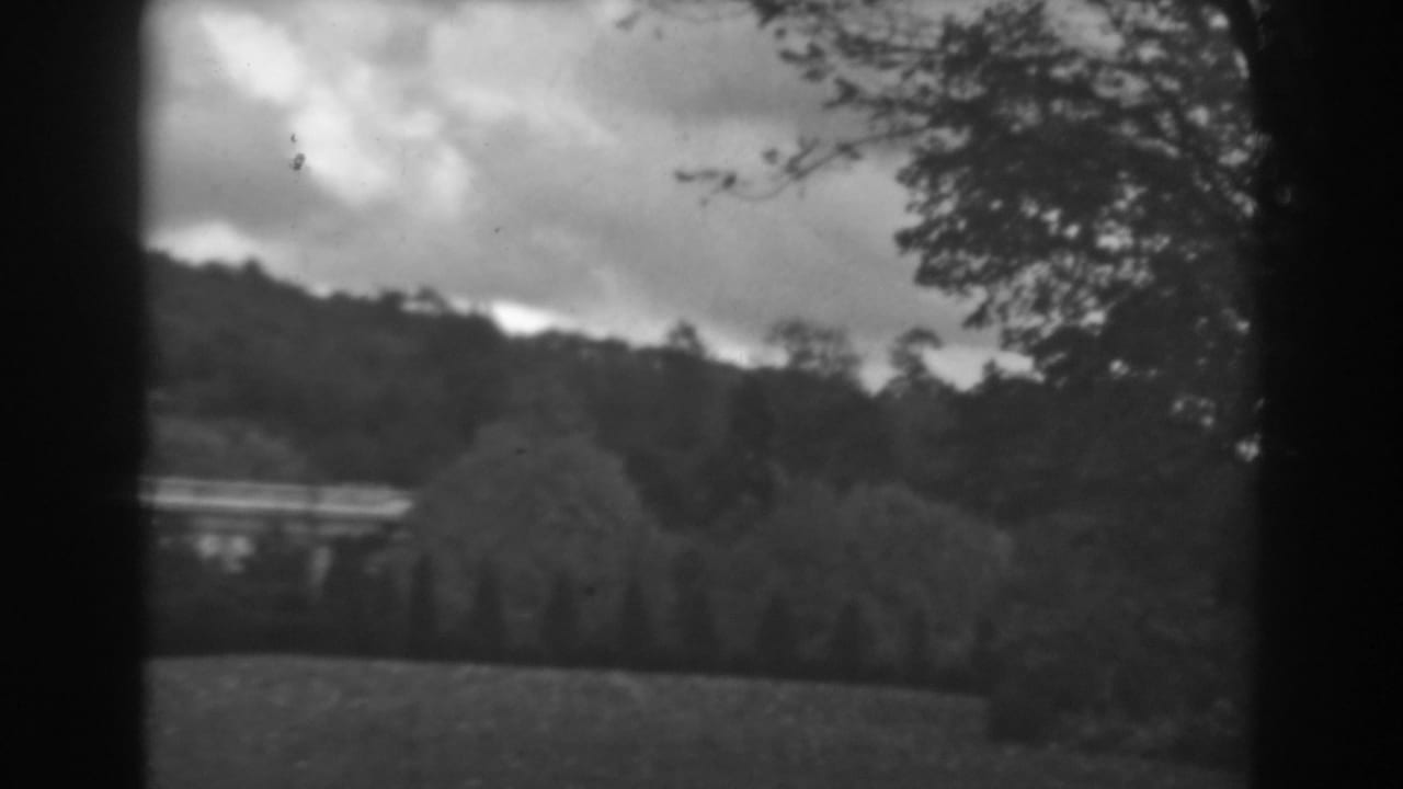 1938 Winchester, Maryland view across grass and trees toward a wooded hillside under heavy clouds, with a pale low structure visible in the distance.