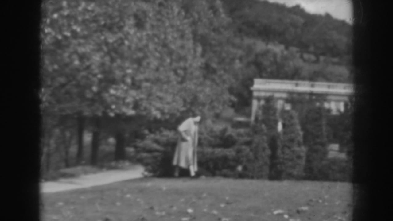 Woman in a long coat walking beside clipped garden shrubs and a lawn.