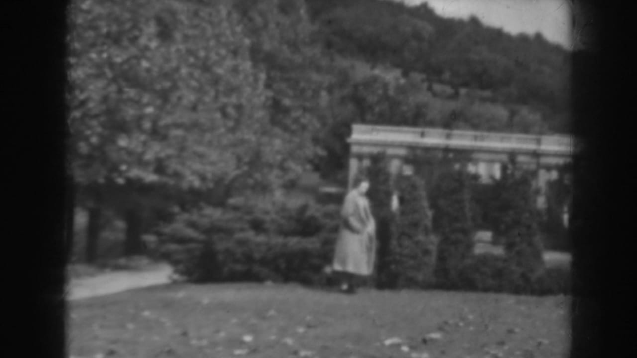 The woman appears near shaped shrubs in a Winchester, Maryland garden, with a path, trees, and a pavilion-like structure in the background.