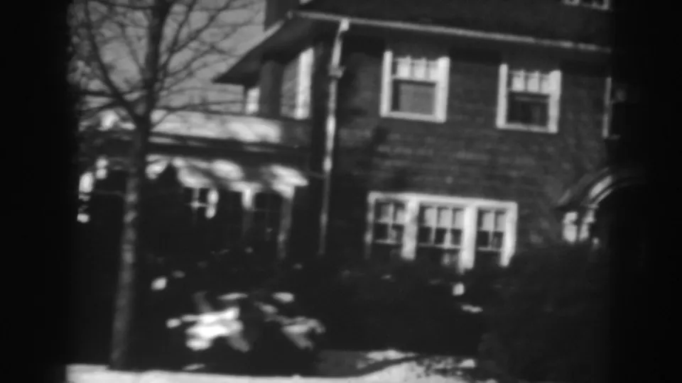 A closer panning frame shows the Winchester residence with upper-story windows, lower window group, bare branches, and yard shrubs.