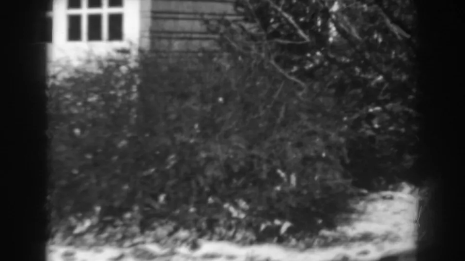 Late shot from the 1938 Winchester home movie showing leafless shrubs, snow on the lawn, and partial house windows.
