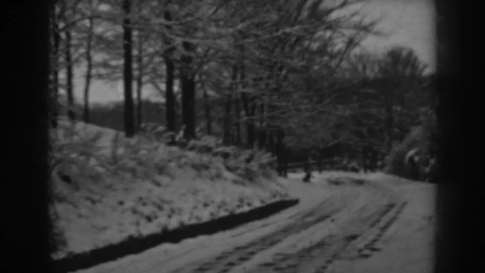 Black-and-white 8mm frame of a winter road curving through a wooded area, with snow on the ground and no people visible.