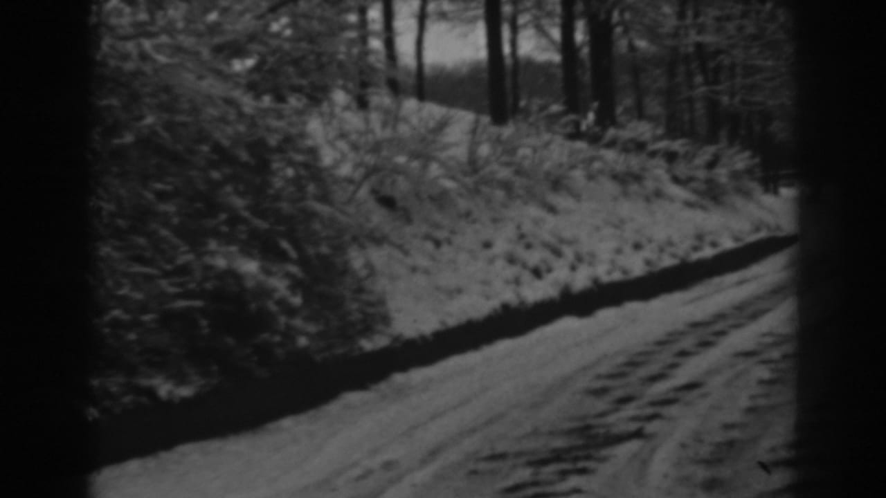 1938 Winchester, Maryland home-movie frame showing a snow-dusted road with tire tracks beside wooded roadside brush.