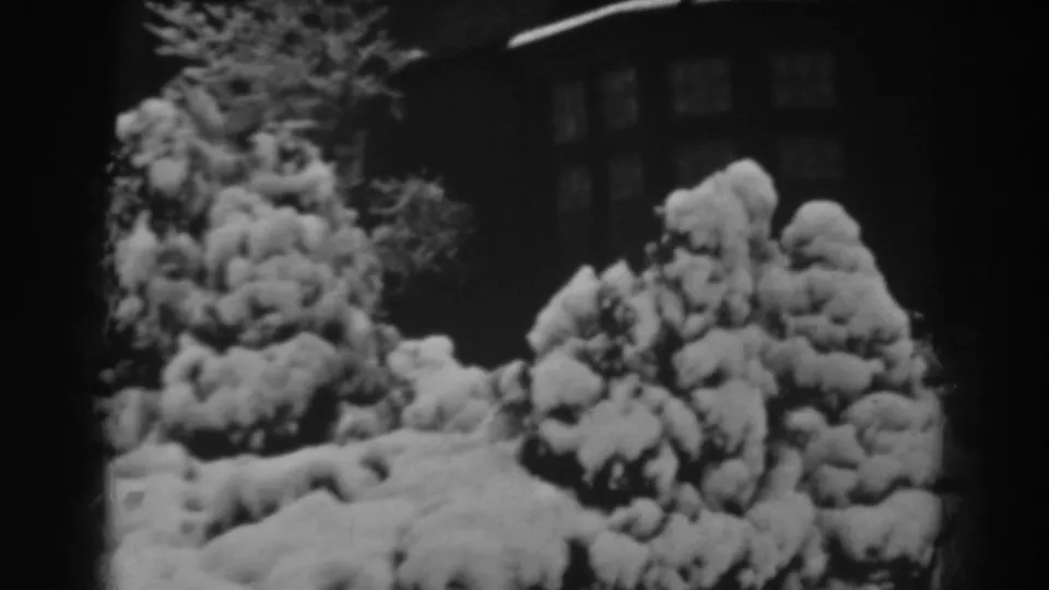 Black-and-white 8mm frame of a snow-covered country yard in Winchester, Maryland, with shrubs and a house behind.