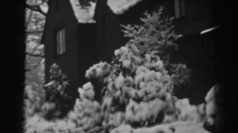 Soft black-and-white 8mm frame showing snow-covered evergreens beside a dark house exterior in Winchester, Maryland.