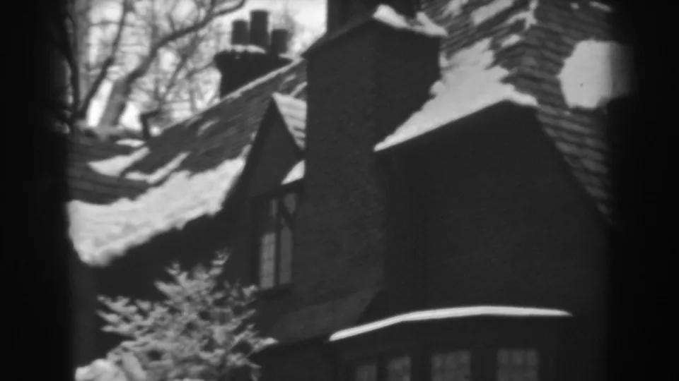 1938 black-and-white 8mm frame of the house exterior, with snow on shingles, a chimney and winter planting below.