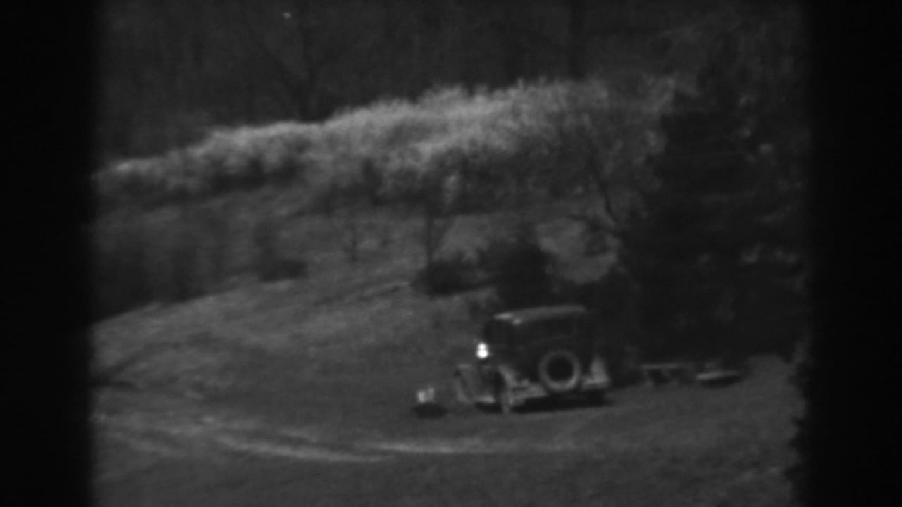 Early automobile on a grassy slope near trees and a small wooden structure.