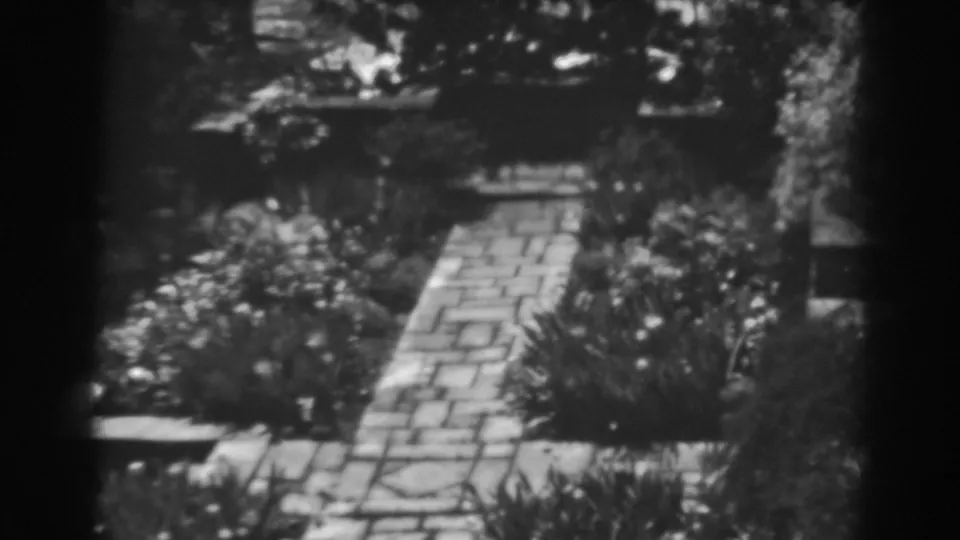 Late-1930s home-movie frame of a cobblestone path receding through a Winchester garden with shrubs and bright sunlight.