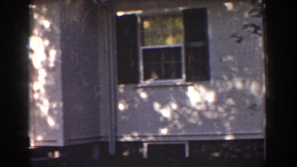 1939 Mount Washington home-movie frame showing a white house facade, dark shutters, dappled tree shadows, and a foreground tree trunk.