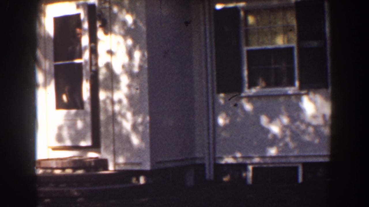 Adult man stands in the open doorway of a white house partly obscured by a tree trunk.