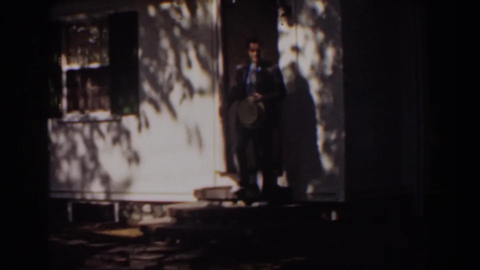 The adult man stands outside the white house on the porch steps, wearing a dark suit and holding a brimmed hat in the 1939 scene.