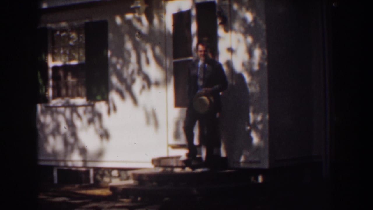 Man in a dark suit stands near a house doorway holding a hat.