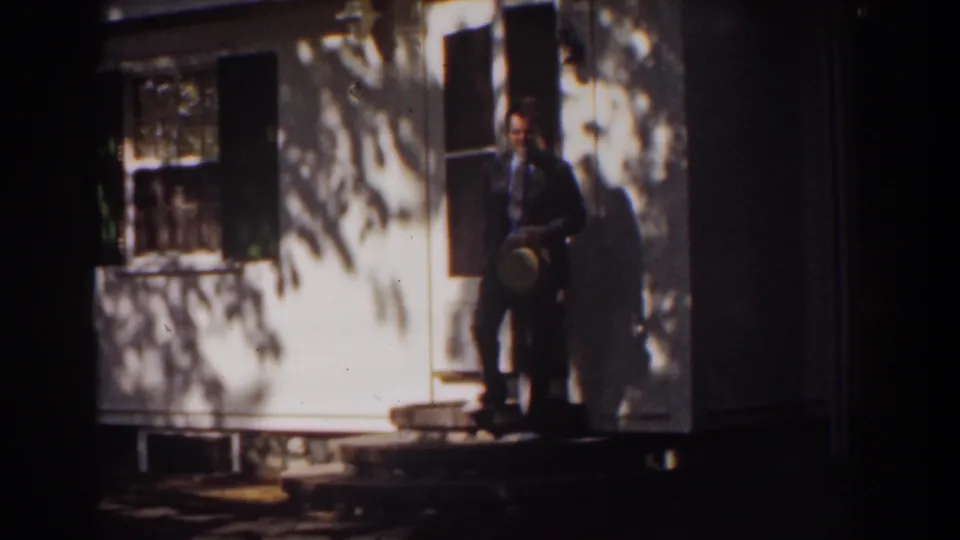 In supplied 1939 Mount Washington home-movie footage, a suited man stands on porch steps holding a hat outside a white house.