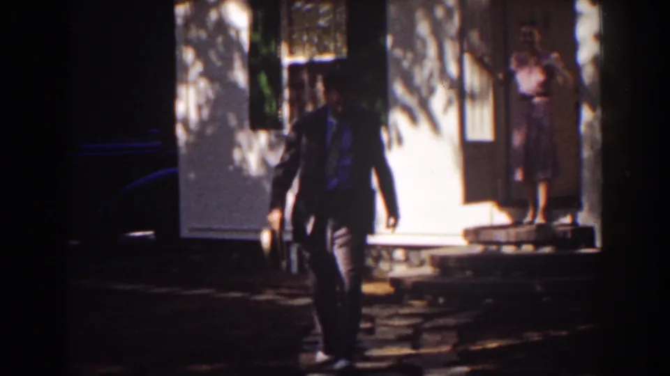 The man walks forward carrying his hat, with the woman still visible in the doorway of the white house behind him.