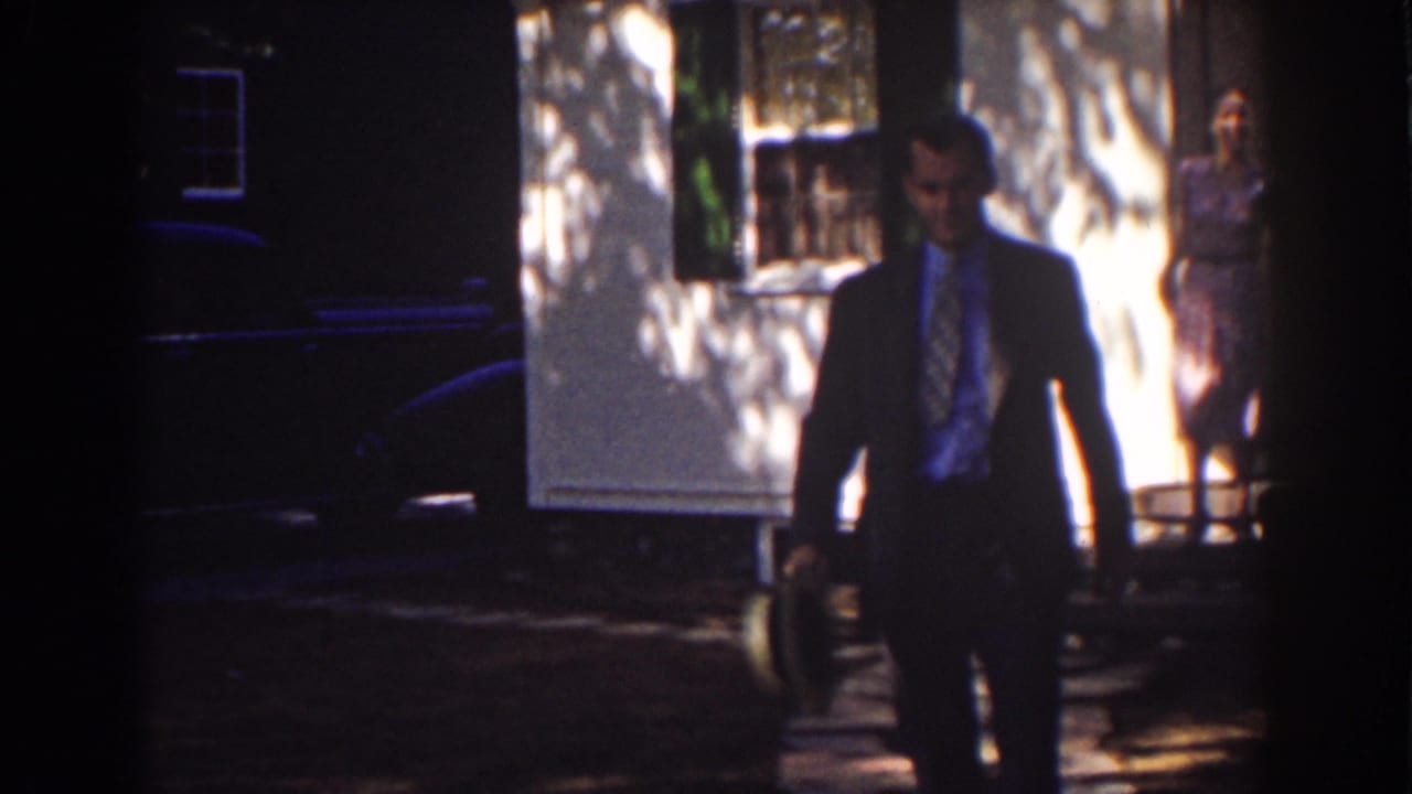 Man in a dark suit walks toward camera as a woman stands near a white house behind him.