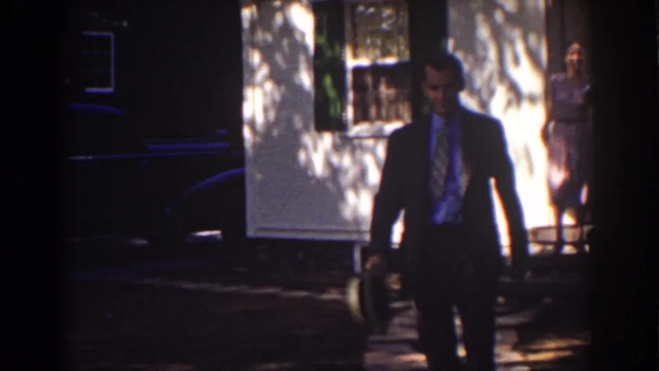 1939 Mount Washington home-movie frame showing a suited man carrying a hat with a woman in a pink dress near a sunlit white house.