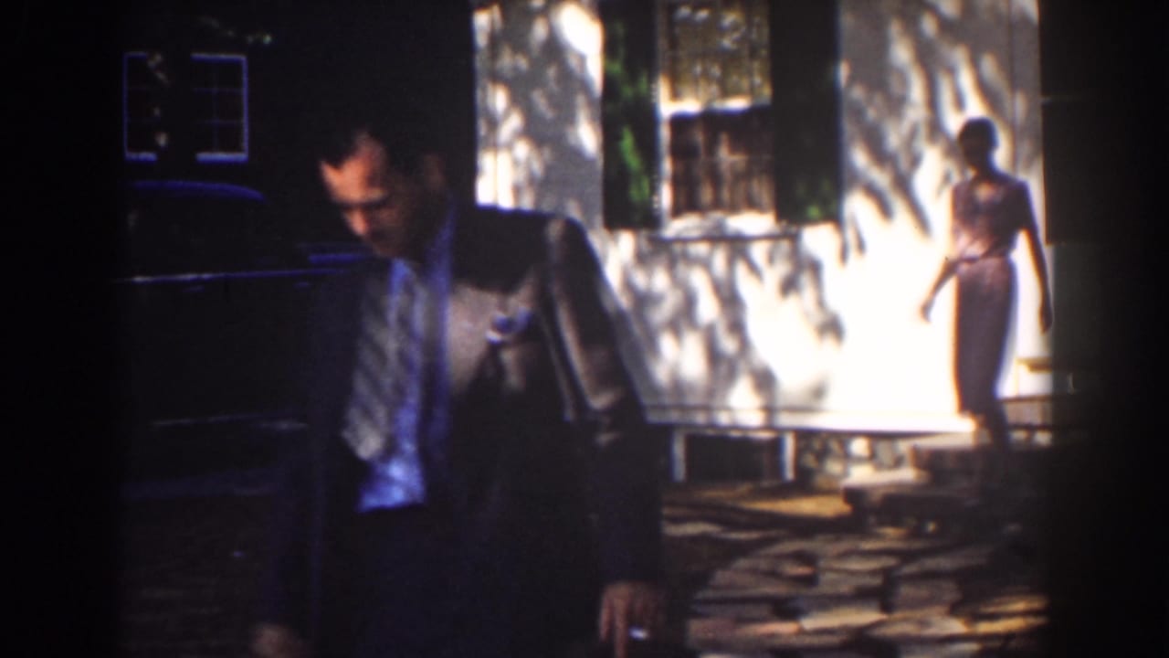 Suited man passes close to the camera while a woman in a pink dress walks near the house.