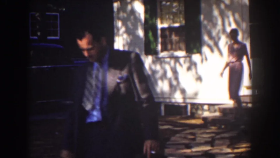 1939 Mount Washington frame with the suited man partly foregrounded and a woman walking along the sunlit house exterior.
