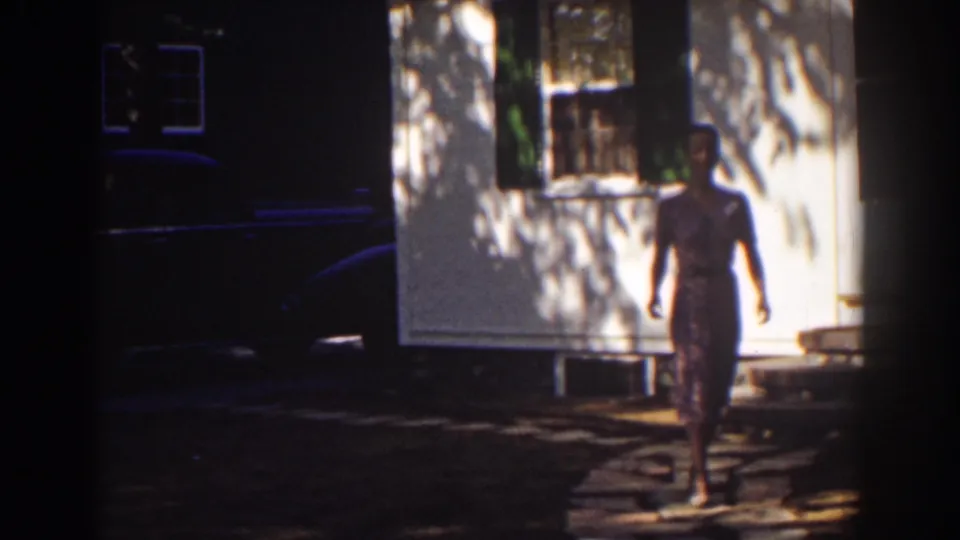 1939 Mount Washington home-movie frame showing a woman in a pink dress approaching along a shaded sidewalk near a white house.
