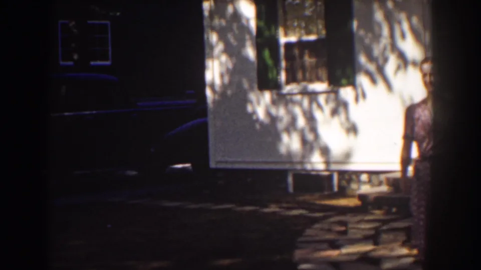 1939 Mount Washington home-movie frame showing a white clapboard house, shaded window, stone path, and a woman partly entering at right.