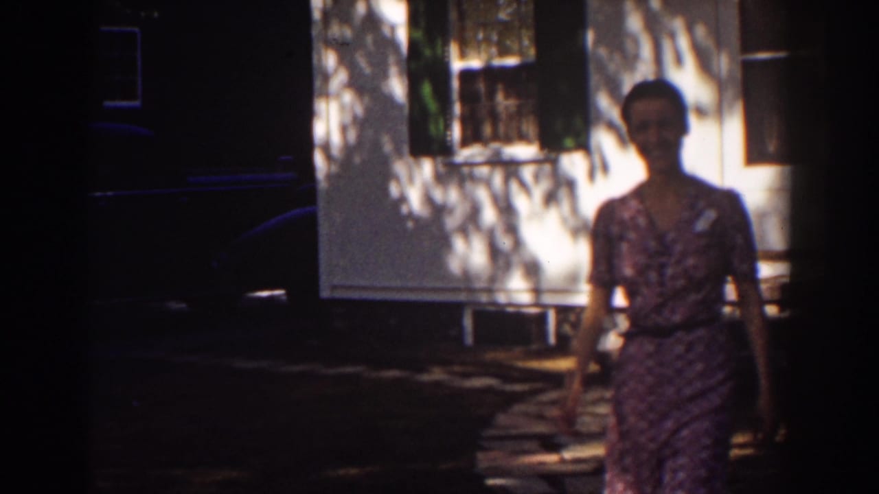 Woman in a patterned dress walking past a white house with shutters and leaf shadows.