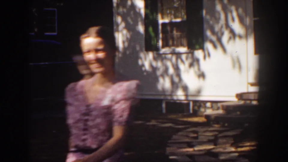 Close home-movie frame of a smiling woman in a patterned dress, blurred by movement, with a white clapboard house and window behind her.