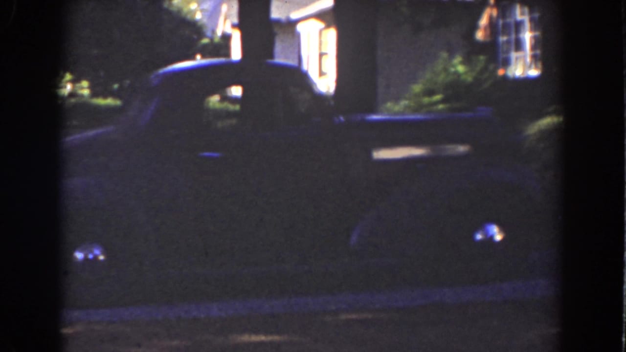 Dark vintage car parked outside a lit residential building at night.