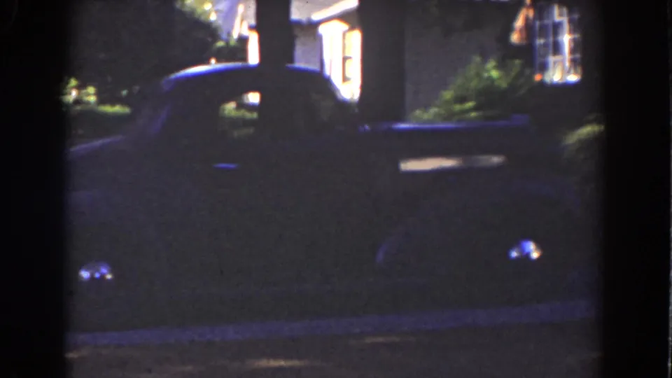 A dark car sits near a curb or driveway outside warm-lit house windows in Mount Washington, New Hampshire, 1939 home-movie footage.