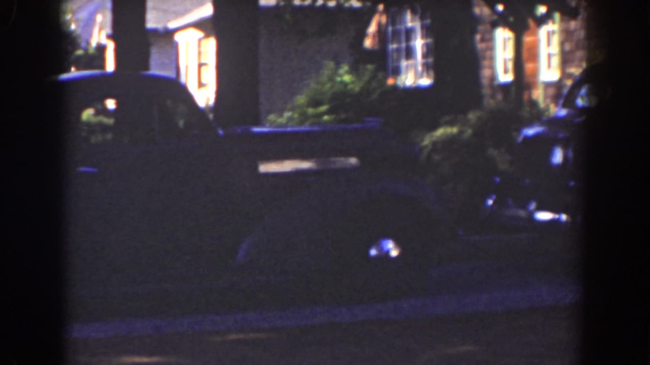Shadowed car beside a residential exterior with illuminated windows.