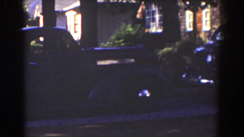 The parked automobile remains mostly in shadow beside shrubs and warm house windows in the 1939 Mount Washington home-movie scene.