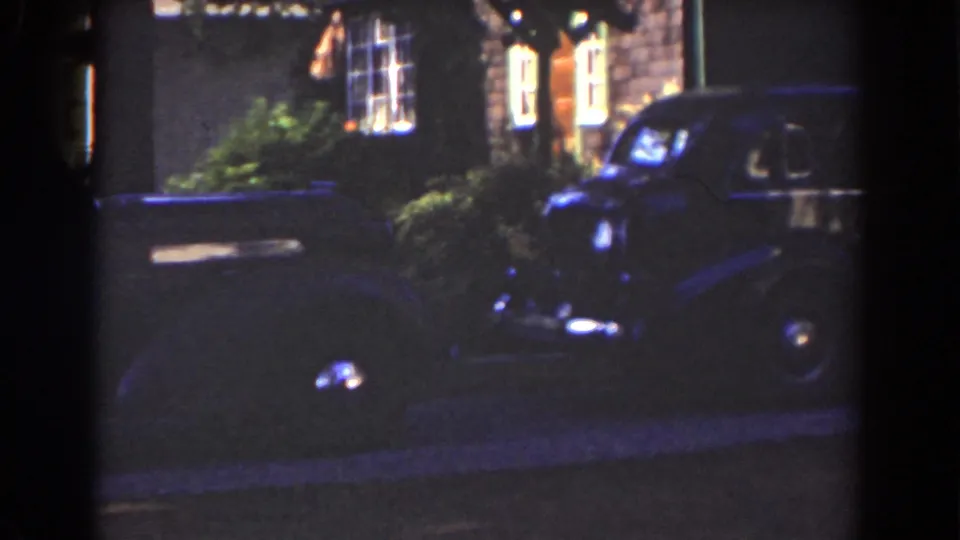 A closer frame shows the car's rounded front, chrome headlamp, and side body outside a lit residential exterior in Mount Washington, 1939.