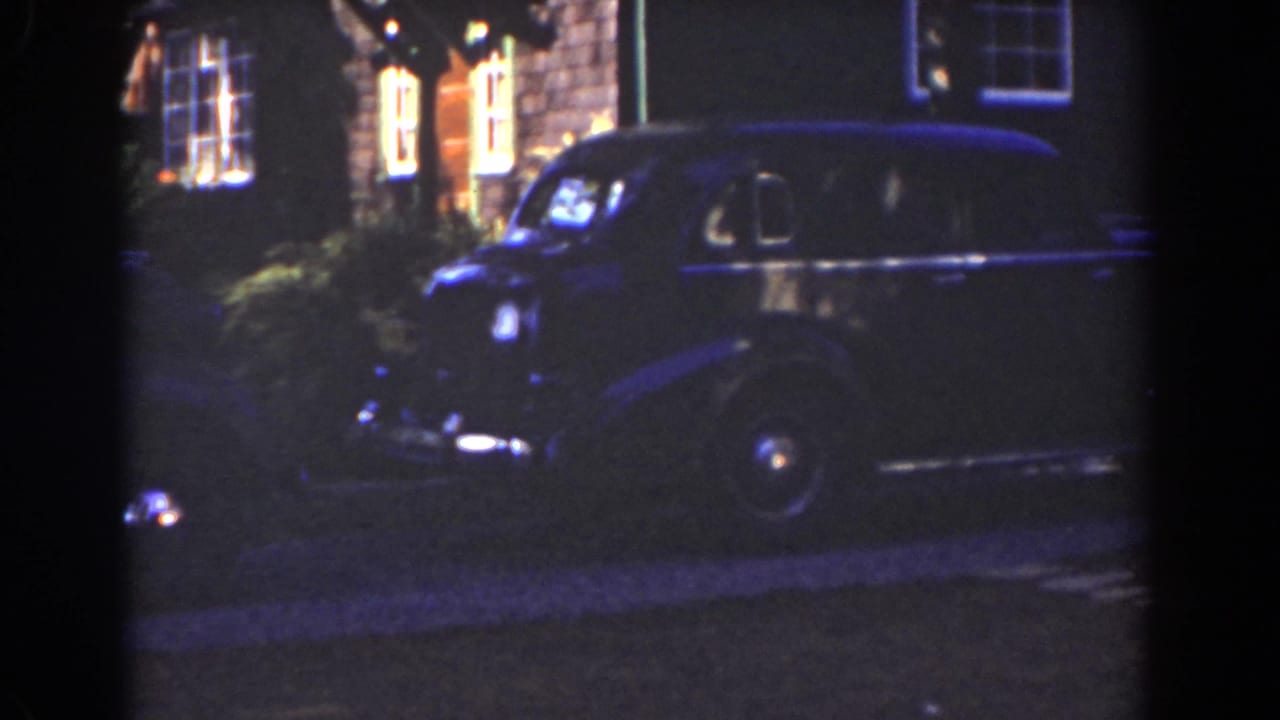 Dark sedan parked beside a house with lit windows at night.
