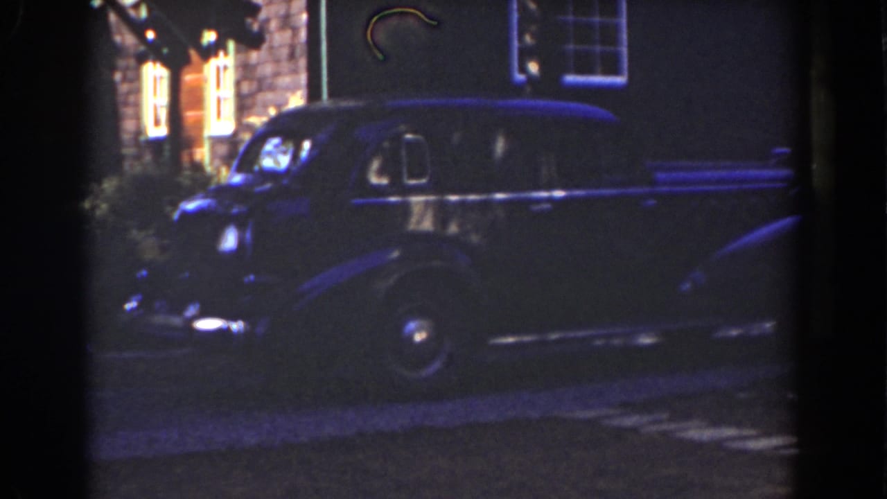 Dark sedan in front of a house with glowing upper windows.