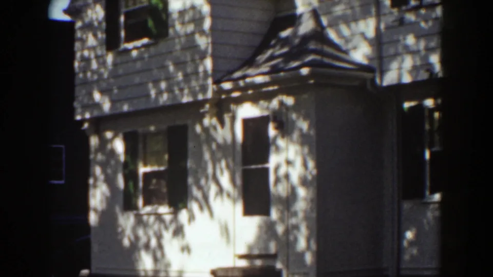 Closer 8mm frame of a white clapboard house with a small gabled entry roof, windows, shutters, and leafy shadows.